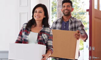 A smiling couple hold moving boxes and enter their new home